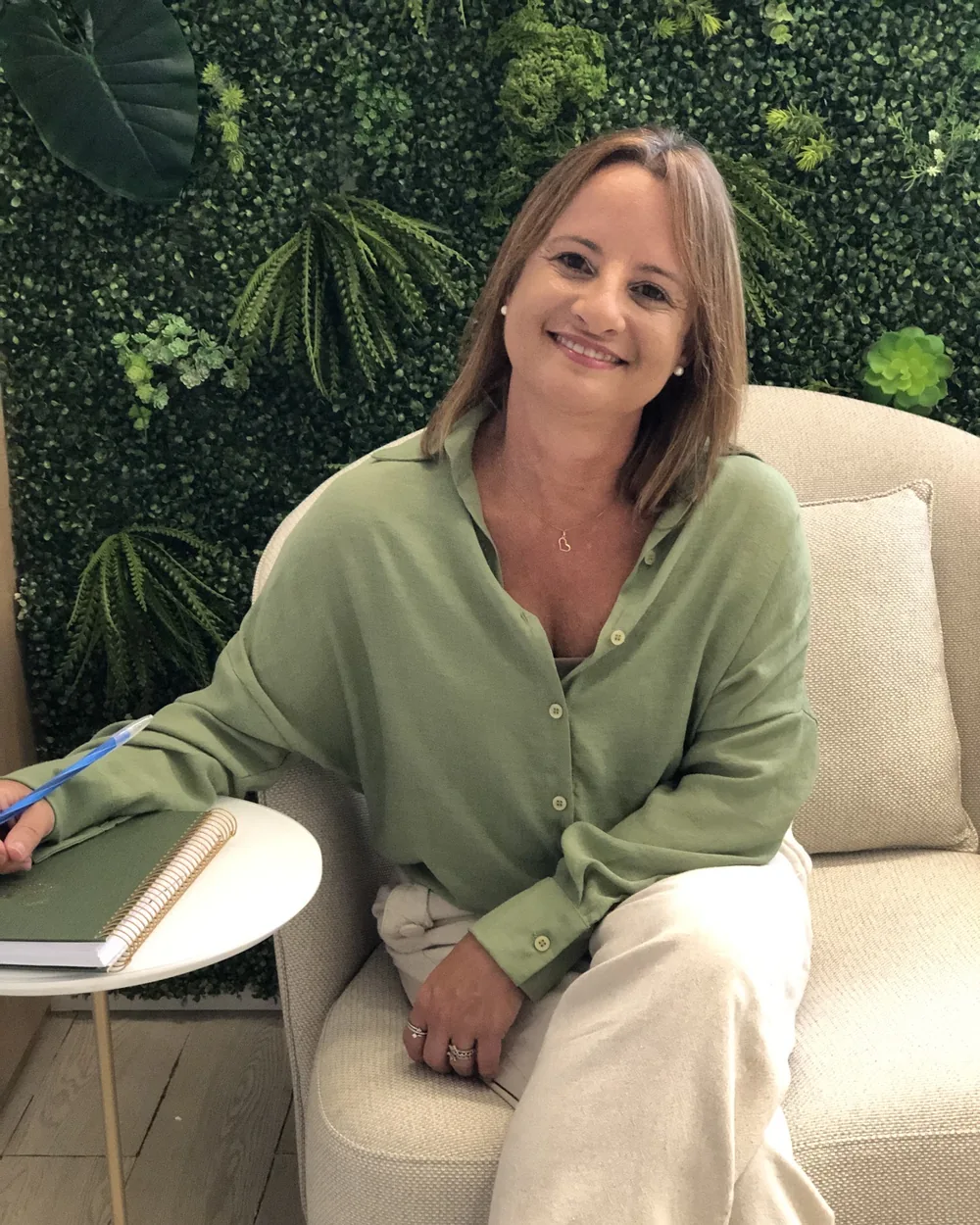 Adriana Assis sorrindo, de camisa verde-oliva, sentada numa poltrona clara do consultório, em frente a uma parede de plantas